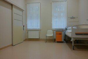 Gulta, krēsls un galds slimnīcas telpā ar logiem un žalūzijām. Rīgas 1. slimnīca.