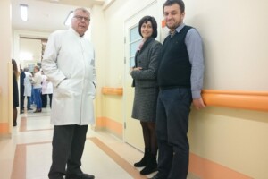 Trīs cilvēki stāv Rīgas 1. slimnīcas koridorā, ģērbušies formālās drēbēs. Fons ar medicīnas personālu.