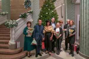 09.12.2021 Gada balva sociālajā nozarē Rīgas 1. slimnīcas darbinieki pie Ziemassvētku egles, turot ziedus un balvas. Fons ar dekorācijām un kāpnēm.