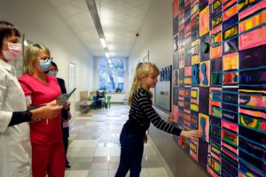 Bērns pieskaras krāsainai sienas gleznai slimnīcas gaitenī, blakus divām medicīnas darbiniecēm.