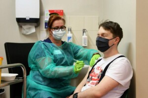 Mediķis vakcinē pacientu Rīgas 1. slimnīcā. Abiem ir maskas, mediķis valkā aizsargtērpu.
