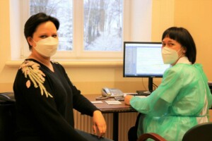 Divas sievietes ar sejas maskām sēž pie galda, blakus dators un telefons. Logā redzams sniegs.