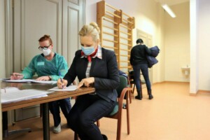 Divas sievietes, viena raksta pie galda, otra stāv. Telpā ir vingrošanas stienis un sanitārā telpa.
