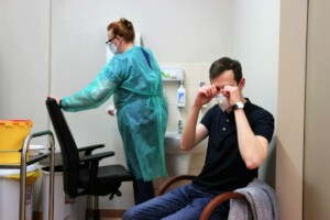 Vīrietis sēž krēslā, pieliekot rokas pie sejas, medicīnas darbinieks strādā fonā.