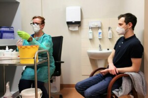 Mediķis ar masku un cimdiem sagatavo injekciju, pacients sēž blakus. Fons: medicīnas telpa.