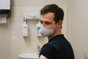 Vīrietis ar masku skatās pār plecu, fona apstākļi medicīnas telpā, redzami dezinfekcijas līdzekļi.