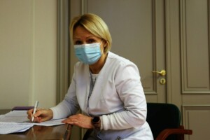 Mediķis ar sejas masku, rakstot pie galda, Rīgas 1. slimnīcas telpās. Papīri un pildspalva uz galda.