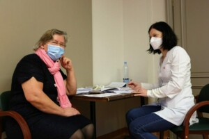 Divas personas, viena ar rozā šalli, otra medicīnas tērpā, sarunājas pie galda Rīgas 1. slimnīcā.
