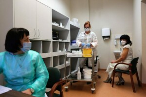 Divas sievietes gaida medicīnisko procedūru, ārste sagatavo materiālus, telpa ir klīniski iekārtota.