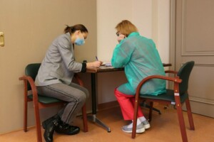 Divas personas pie galda, viena raksta, otra klausās. Abas valkā maskas, viena ir medicīniskā apģērbā.