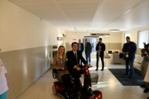 Divi cilvēki sēž uz elektriskā skūtera Rīgas 1. slimnīcas gaitenī, apkārt citi cilvēki un fotogrāfi.