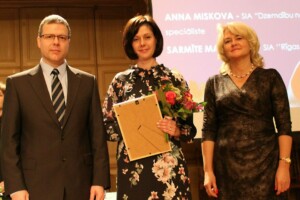Trīs cilvēki uz skatuves, Anna Miskova ar ziediem un diplomu, fons ar tekstu "INOVATĪVAS". Rīgas 1. slimnīca.
