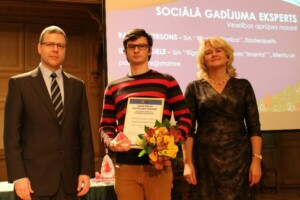 Divi vīrieši un sieviete stāv pie ekrāna, Ravis Pētersons tur balvu par sociālo darbu. Fons: "Sociālā gadījuma