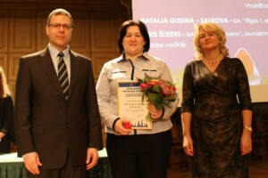 Attēlā ir trīs cilvēki, no kuriem vidū ir Nataļja Gudina-Salkova ar balvu "Gada balva sociālajā nozarē".
