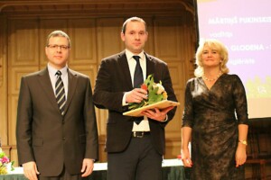 Trīs cilvēki uz skatuves, viens ar ziediem un diplomu. Fons ar tekstu "Mārtiņš Pukinskis, vadītājs".