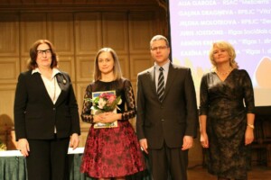 Četri cilvēki stāv pie galda, divi ar ziediem un diplomiem, Rīgas 1. slimnīcas pasākumā.