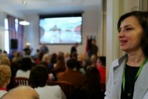 edf Rīgas 1. slimnīcas pasākums ar auditoriju un prezentāciju ekrānā. Sieviete baltā apģērbā stāv priekšplānā.
