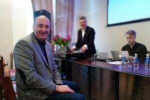 edf Vīrietis ar pelēku uzvalku sēž pie galda, fonā citi cilvēki un ziedi. Rīgas 1. slimnīcas pasākums.