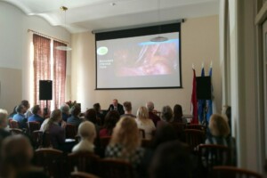 edf Konference Rīgas 1. slimnīcā, runātājs pie galda, ekrānā attēls ar tekstu "Bokovaya stenka taza".