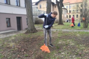 edf Jauns vīrietis ar grābekli sakopj zālienu Rīgas 1. slimnīcas teritorijā. Fons ar ēkām un citiem cilvēkiem.