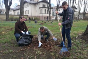 edf Trīs vīrieši sakopj lapas Rīgas 1. slimnīcas teritorijā, izmantojot maisu un grābekli.