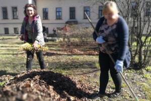edf Divas sievietes dārzā, viena tur dzeltenu grābekli, otra ar nūju, apkārt lapas un zāle. Rīgas 1. slimnīca.