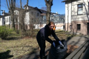 edf Sieviete savāc atkritumus melnā maisā Rīgas 1. slimnīcas teritorijā, fonā redzamas ēkas un koki.