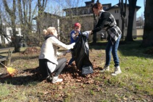 edf Trīs cilvēki sakopj lapas parkā, izmantojot melnu maisu un rīkus. Rīgas 1. slimnīcas apkārtnē.