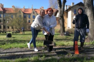 edf Trīs sievietes dārzā, viena ar lāpstu, otra ar slotu, fons - Rīgas 1. slimnīcas ēka un zaļa zāle.