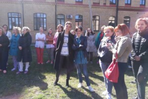 dav Grupas cilvēku stāv Rīgas 1. slimnīcas pagalmā, saulainā dienā, daži runā un skatās uz telefonu.