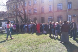 dav Grupa cilvēku stāv zālē, klausoties runātāju Rīgas 1. slimnīcas pagalmā saulainā dienā.