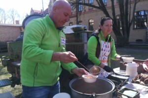 dav Divi cilvēki zaļās jakās pasniedz zupu ārpus Rīgas 1. slimnīcas. Redzami trauki un pārtikas produkti.
