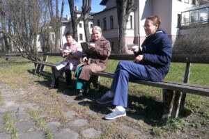 dav Trīs sievietes sēž uz soliņa, baudot ēdienu Rīgas 1. slimnīcas dārzā saulainā dienā.