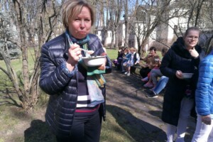 dav Sieviete ar bļodu ēdiena, stāvot parkā, apkārt citi cilvēki, kas sēž uz soliņa. Rīgas 1. slimnīca.