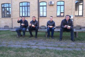 dav Četri vīrieši sēž uz sola, ēdot, Rīgas 1. slimnīcas pagalmā. Divi no viņiem rāda mierinājuma žestu.