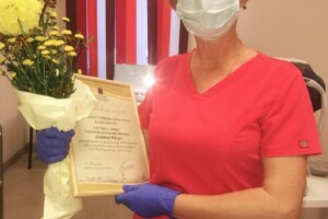 dav Sieviete rozā medicīnas tērpā ar ziediem un atzinības rakstu no Rīgas 1. slimnīcas.