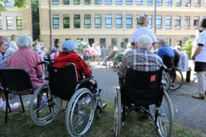 Attēlā redzami seniori ratiņkrēslos, piedaloties pasākumā Rīgas 1. slimnīcā. Fons ar ziediem un cilvēkiem.