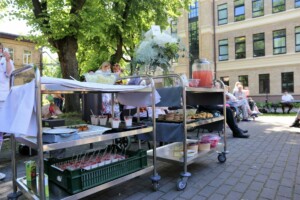 Pasākums Rīgas 1. slimnīcā, galds ar uzkodām, dzērieniem un ziediem, apmeklētāji sēž uz soliņiem.