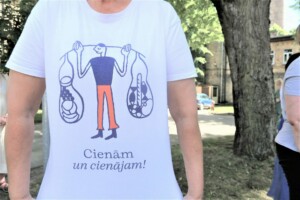 Attēlā redzams cilvēks ar T-kreklu, uz kura ir uzraksts "Cienām un cienājam!" un zīmējums ar svaru.