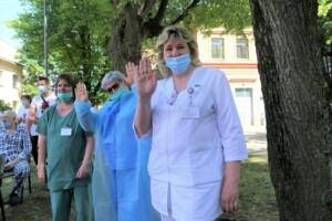 Trīs mediķi un divi pacienti sveicina no Rīgas 1. slimnīcas dārza, visi valkā maskas.