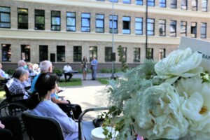 Pasākums Rīgas 1. slimnīcas pagalmā, apmeklētāji sēž krēslos, ziedi priekšplānā, uz skatuves divi izpildītāji.