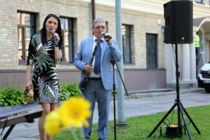 Divi izpildītāji uzstājas ārā, viens ar mikrofonu, otrs ar klarinetu, fons ar zālēm un ēku.
