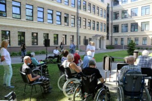 Pasākums Rīgas 1. slimnīcas pagalmā, cilvēki ratiņkrēslos klausās uzstāšanos.