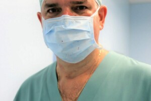 Ārsts ar masku un cepuri, tērpies zaļā medicīnas apģērbā, stāv pie sienas Rīgas 1. slimnīcā.
