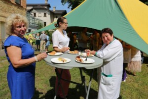 Divas sievietes un viena medicīnas darbiniece ēd un sarunājas zem teltī. Fons: cilvēki un ēdiens uz galda.
