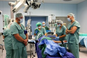 Ķirurģijas telpa ar medicīnas personālu, kas strādā pie pacienta operācijas. Redzami medicīnas instrumenti.