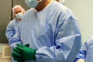 Ārsti operāciju zālē, ģērbušies medicīniskajā apģērbā un aizsargbrillēs, gatavojas procedūrai.