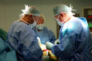 Trīs ķirurgi veic operāciju, valkājot medicīniskos apģērbus un maskas, slimnīcas telpā.