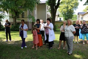Rīgas 1. slimnīcas darbinieki un apmeklētāji stāv dārzā, svinot pasākumu ar ziedu vainagu.