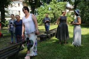 Grupas cilvēku tikšanās Rīgas 1. slimnīcas dārzā, saulaina diena, dažādas drēbes un apģērbu stili.